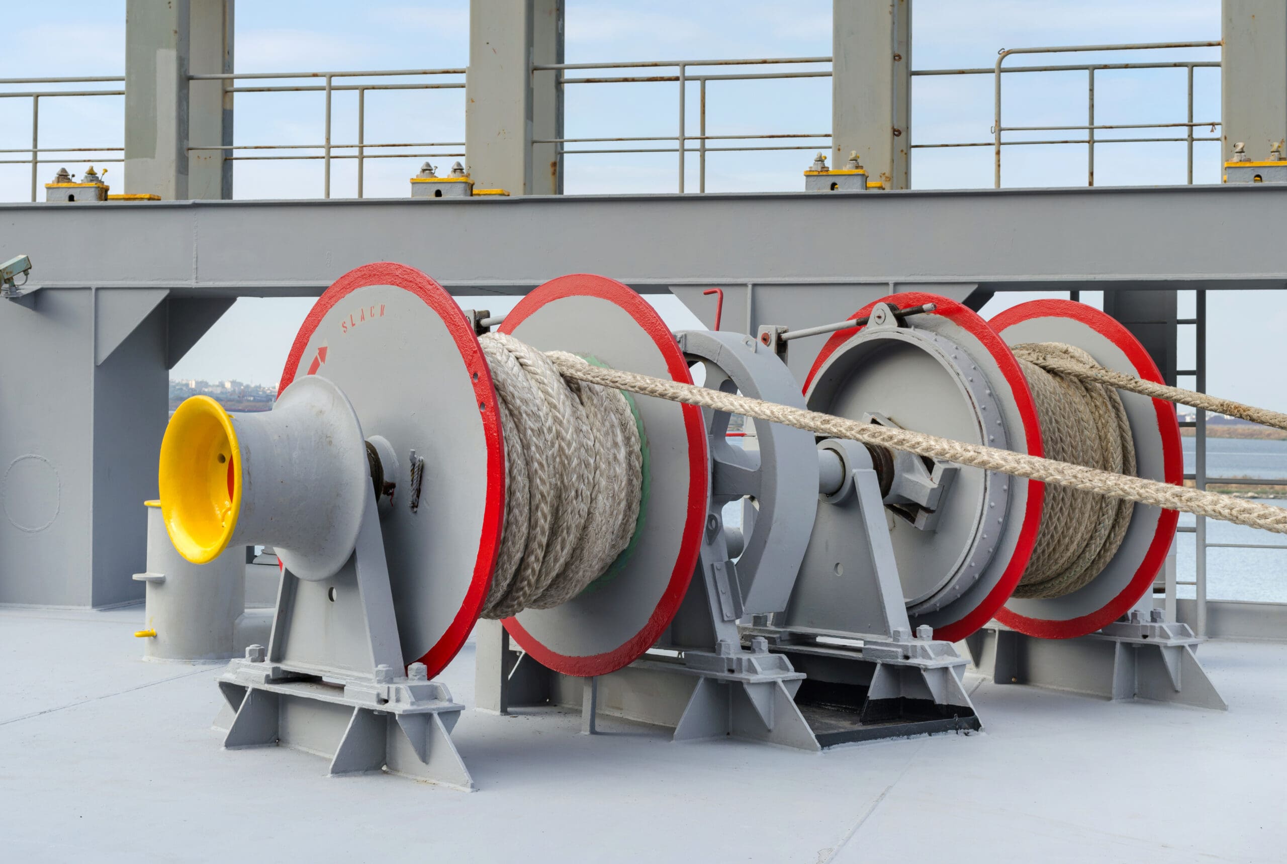 Three large industrial rope winches with thick ropes wound around them are mounted on the deck of a ship. The winches have gray bodies with red accents and are positioned near the ship’s railing.