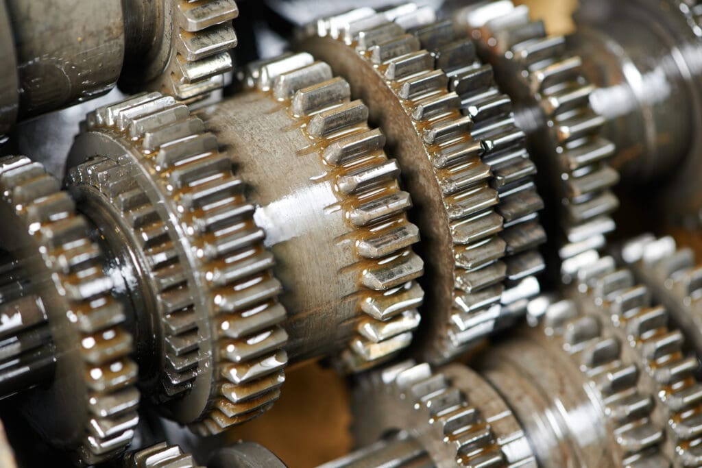 Close-up view of several interlocking, metal gears covered in oil, showing shiny, toothed edges and an industrial, mechanical setting.