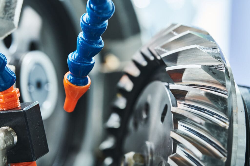 Close-up of a metal gear being machined, with blue and orange coolant hoses directing fluid onto the gear’s shiny, angular teeth. The background is blurred, emphasizing the precision of the industrial process.