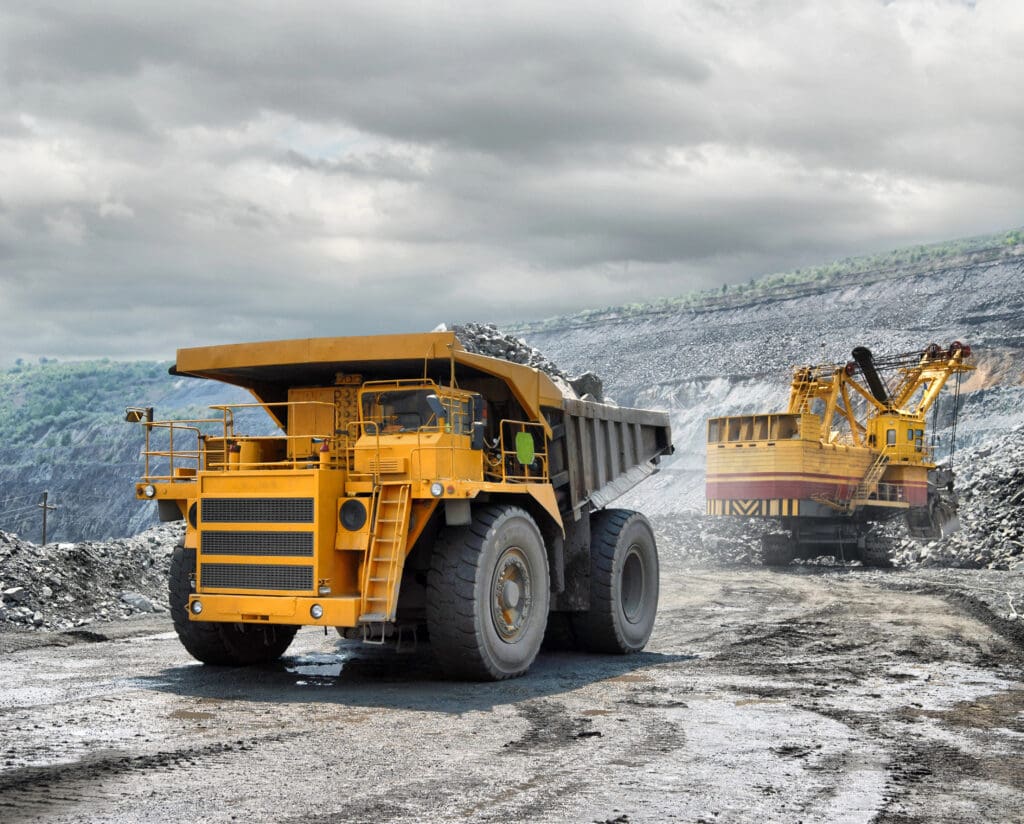 A large yellow dump truck loaded with rocks drives through an open-pit mining site, while an excavator loads material in the background under a cloudy sky.
