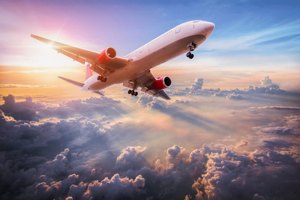 A commercial airplane with red engines flies above clouds at sunset, with sun rays streaming through the sky and illuminating the clouds below.