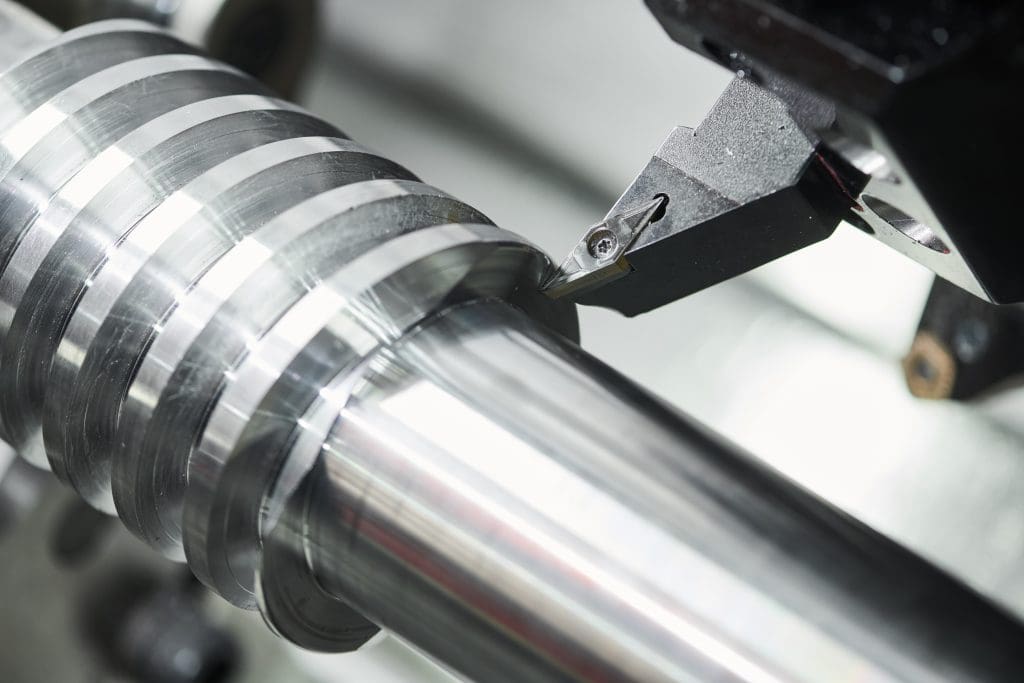 Close-up of a metalworking lathe cutting tool shaping a shiny, cylindrical metal piece with grooves. The tool is in contact with the metal, creating precise, smooth surfaces.