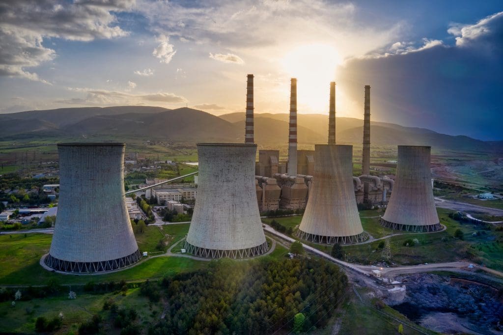 Aerial view of a power plant with four large cooling towers and several tall chimneys, set against mountains at sunset, surrounded by greenery and industrial buildings.