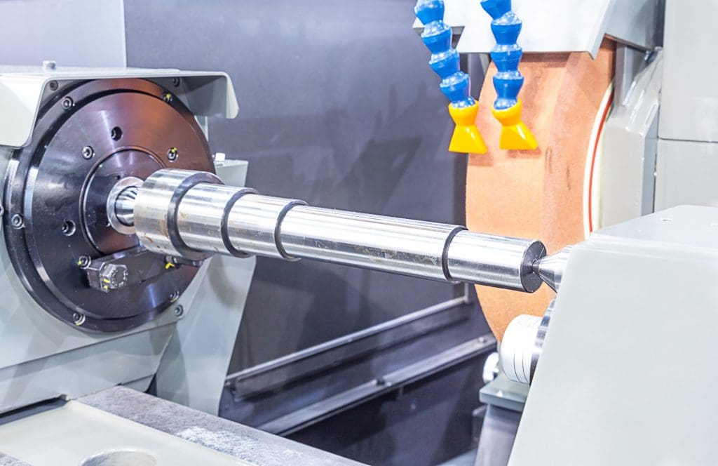 A metal cylindrical shaft is being precisely ground on a CNC grinding machine, with coolant hoses positioned nearby and an orange grinding wheel in the background.