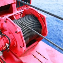 A close-up of a red marine winch with black steel cable on a ship’s deck, with the blue ocean visible in the background.