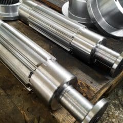 Two large, shiny metal machine shafts with gear-like ridges are lying parallel on a wooden surface in an industrial setting. Circular metal components are partially visible in the background.