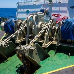 A close-up view of a ship's anchor windlass system with two large chains and covered winches on the deck, with the ocean and part of the ship's structure visible in the background.