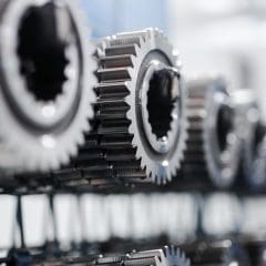 Close-up view of several shiny metal gears lined up in a row, with a shallow depth of field creating a blurred background. The gears have interlocking teeth and are used in mechanical machinery.