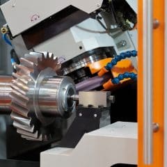 A close-up of an industrial CNC machine grinding a large metal gear with sharp angled teeth. Blue coolant tubes and orange guards surround the rotating gear on the workbench.