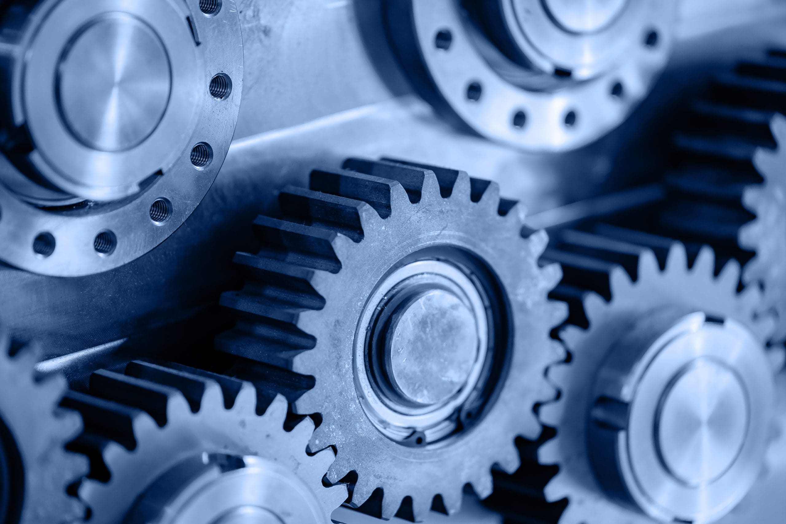 Close-up view of several interlocking metal gears with a blue tint, highlighting their teeth and circular shapes, suggesting machinery or mechanical engineering concepts.