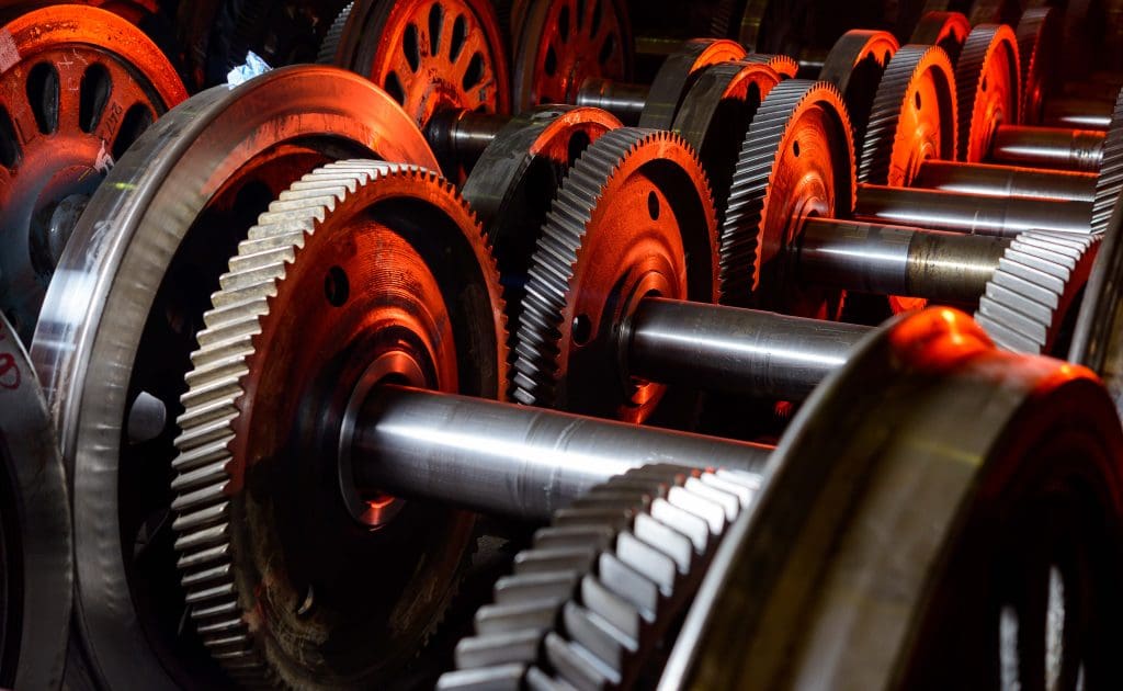 A close-up view of several large metal gears and shafts in a machine, lit by an orange-red light that highlights their shiny, intricate surfaces.