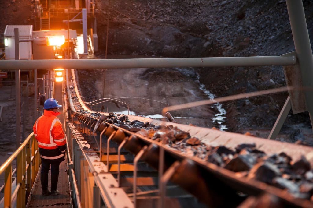 A worker in a blue helmet and orange safety jacket observes a conveyor belt transporting rocks at an industrial mining site, illuminated by orange lights during dusk or dawn.