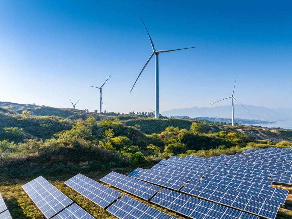 Solar panels and wind turbines on a grassy hillside under a clear blue sky, with mountains visible in the distance. The image highlights renewable energy sources in a natural landscape.