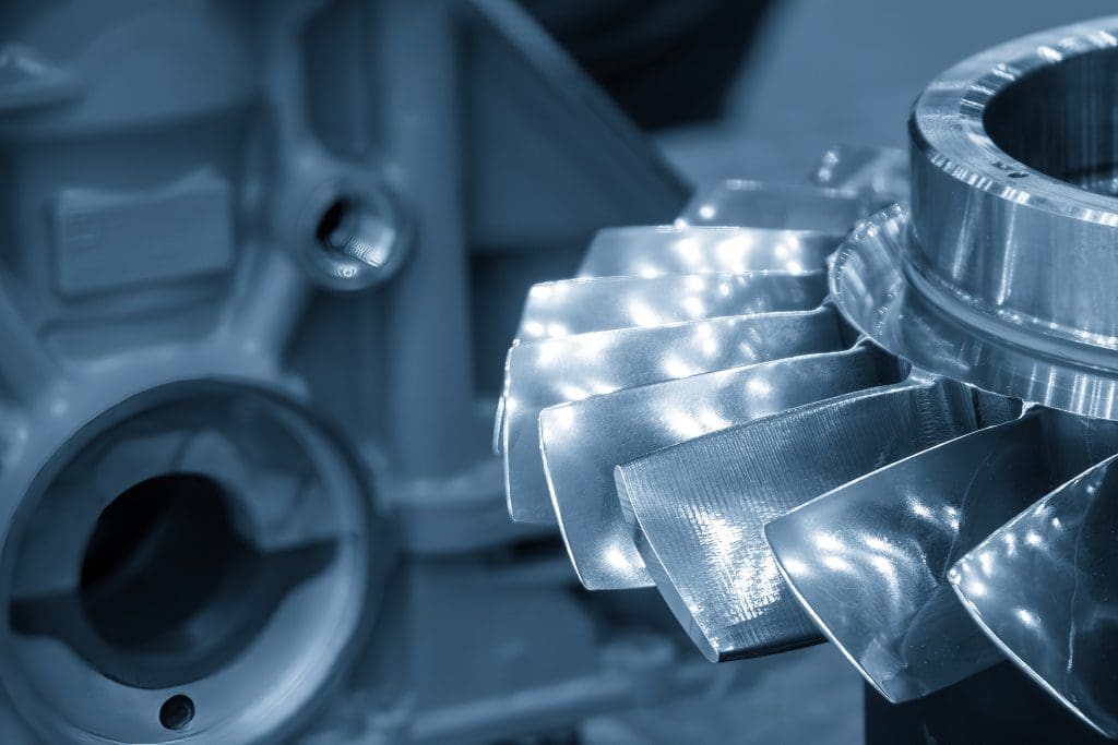 Close-up of a shiny metal turbine or compressor blade assembly, showing smooth, reflective blades with a blurred industrial machine part in the background. The image has a cool blue tone.
