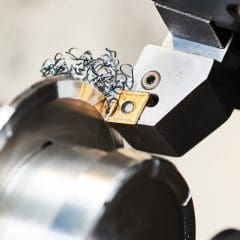 A metal lathe cutting tool shapes a shiny cylindrical metal workpiece, producing spiraled metal shavings during the machining process.