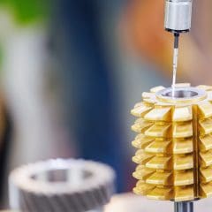 A close-up of a gear-cutting tool being measured with a precision probe. The gold-colored cutting edges are arranged in a helical pattern, and the background is blurred with green and blue tones.