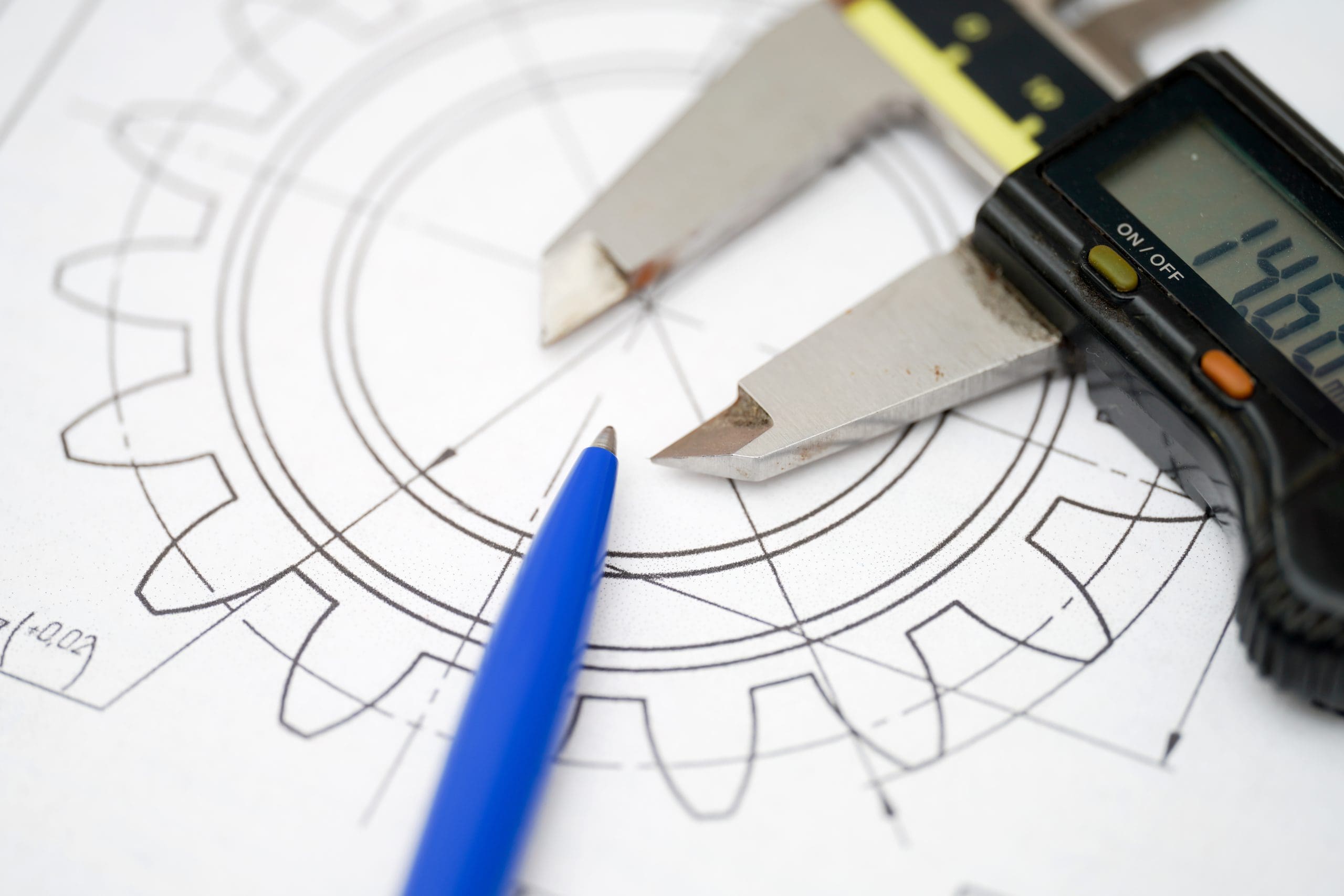 A close-up of a digital caliper and a blue pen resting on a technical drawing of a gear, illustrating precision engineering and mechanical design.