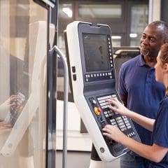 A woman operates an industrial control panel with multiple buttons and screens, while a man stands beside her, watching and providing guidance. They are in a modern industrial or manufacturing setting.