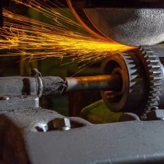 A close-up of a gear shaft being ground by a rotating abrasive wheel, with bright orange sparks flying from the point of contact.