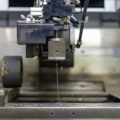 Close-up of a wire-cut electrical discharge machine (EDM) in operation, with a thin metal wire precisely cutting through a piece of metal. The machine is surrounded by metallic components.