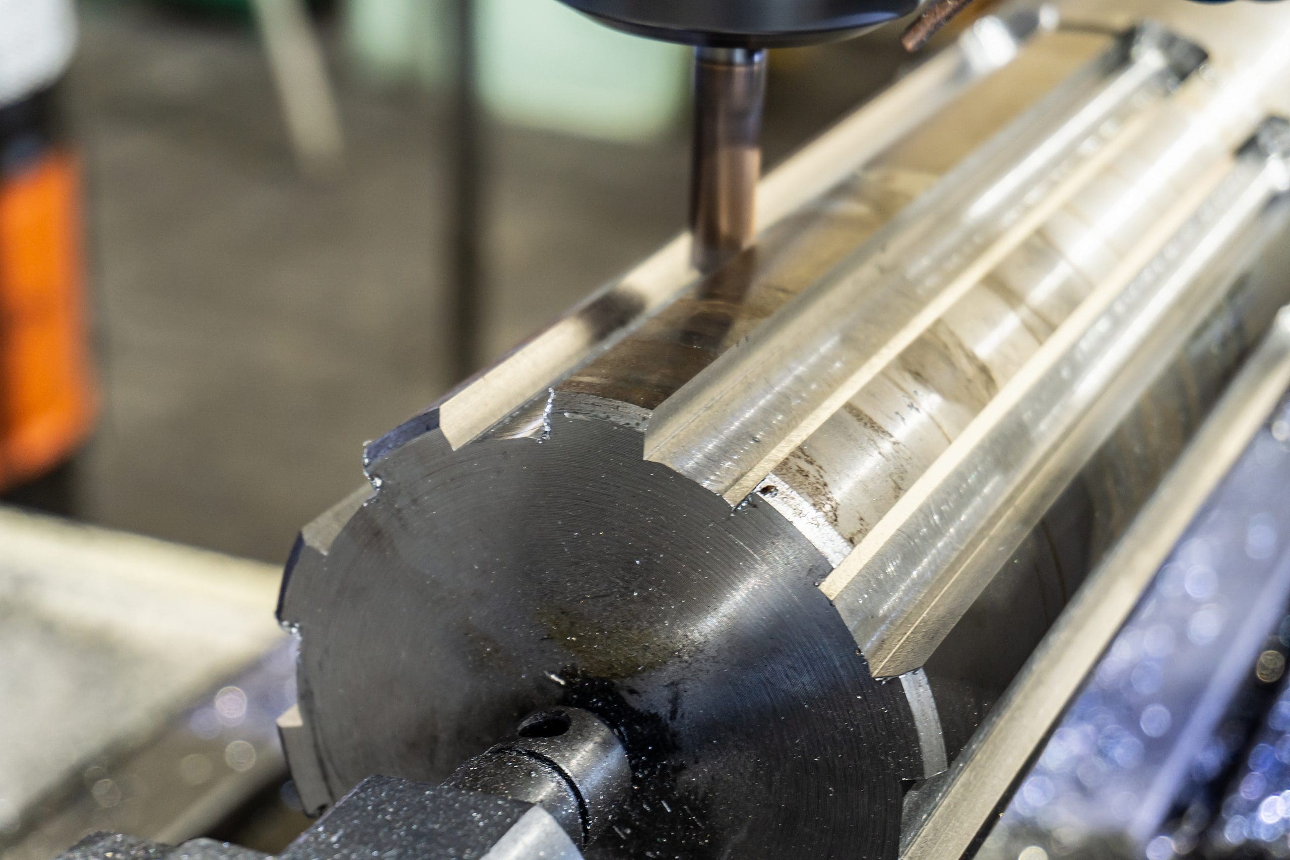 A close-up of a cylindrical metal workpiece being machined on a milling machine, showing precise grooves and shiny surfaces created by the cutting tool.