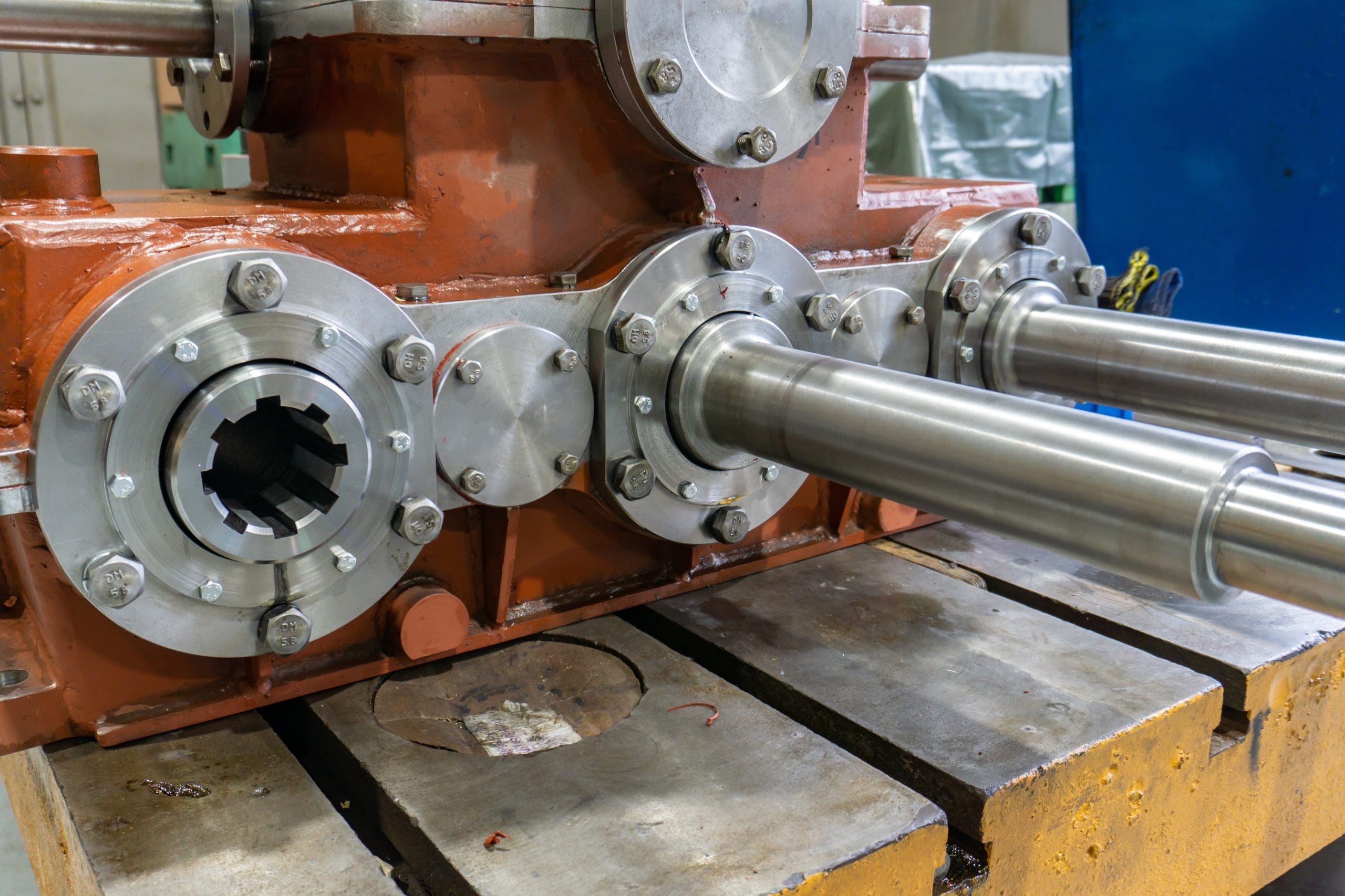 Close-up of a heavy-duty industrial gearbox with two large steel shafts extending outward, mounted on a yellow metal base in a workshop setting. The gearbox has a rust-orange and metallic finish.