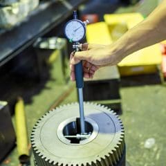 A person’s hand uses a bore gauge to measure the internal diameter of a large metal gear in an industrial workshop setting.
