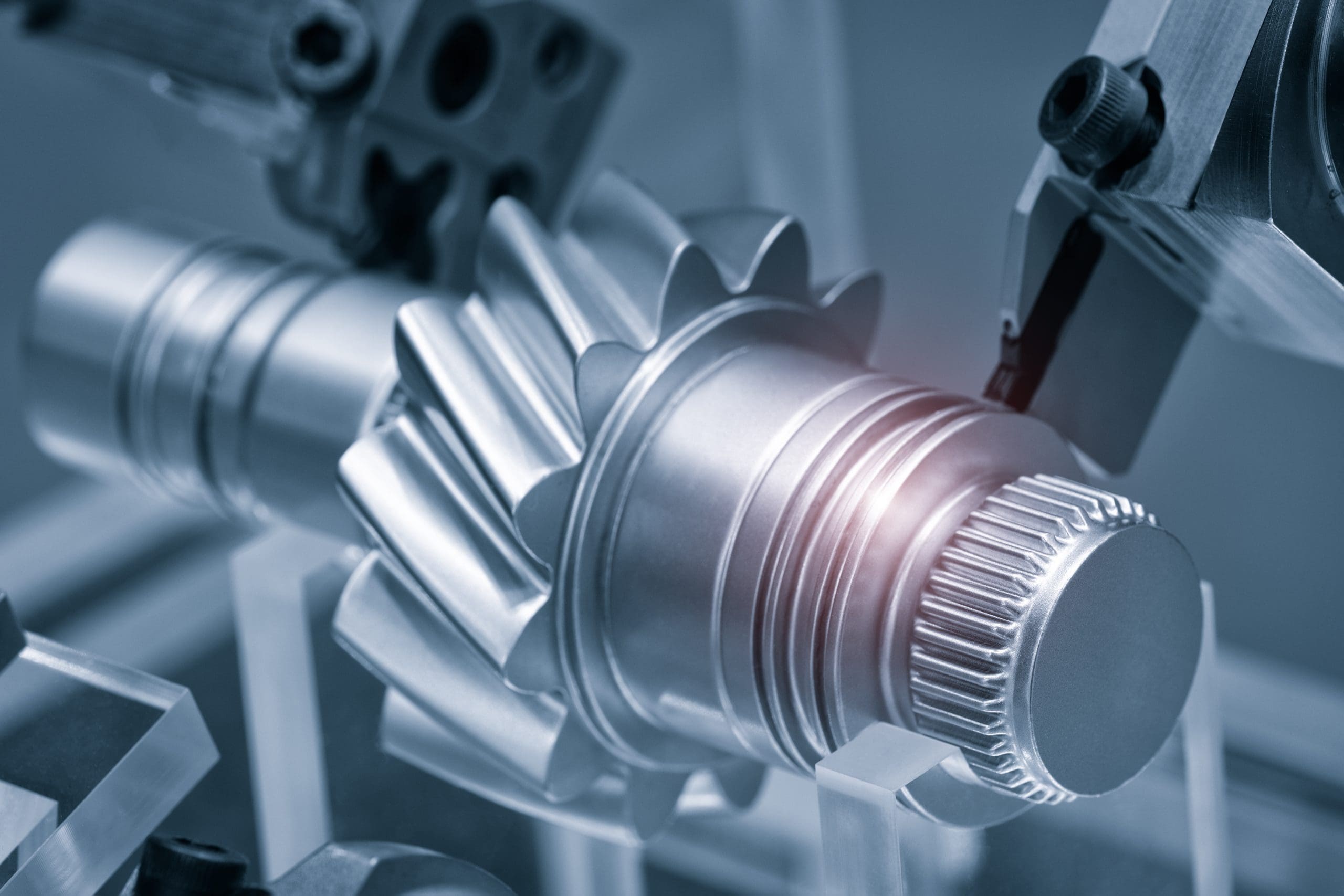 Close-up view of a shiny metal gear being machined by CNC equipment, with precise tools shaping the gear’s teeth and surfaces. The image has a cool blue tone, highlighting the industrial and mechanical setting.