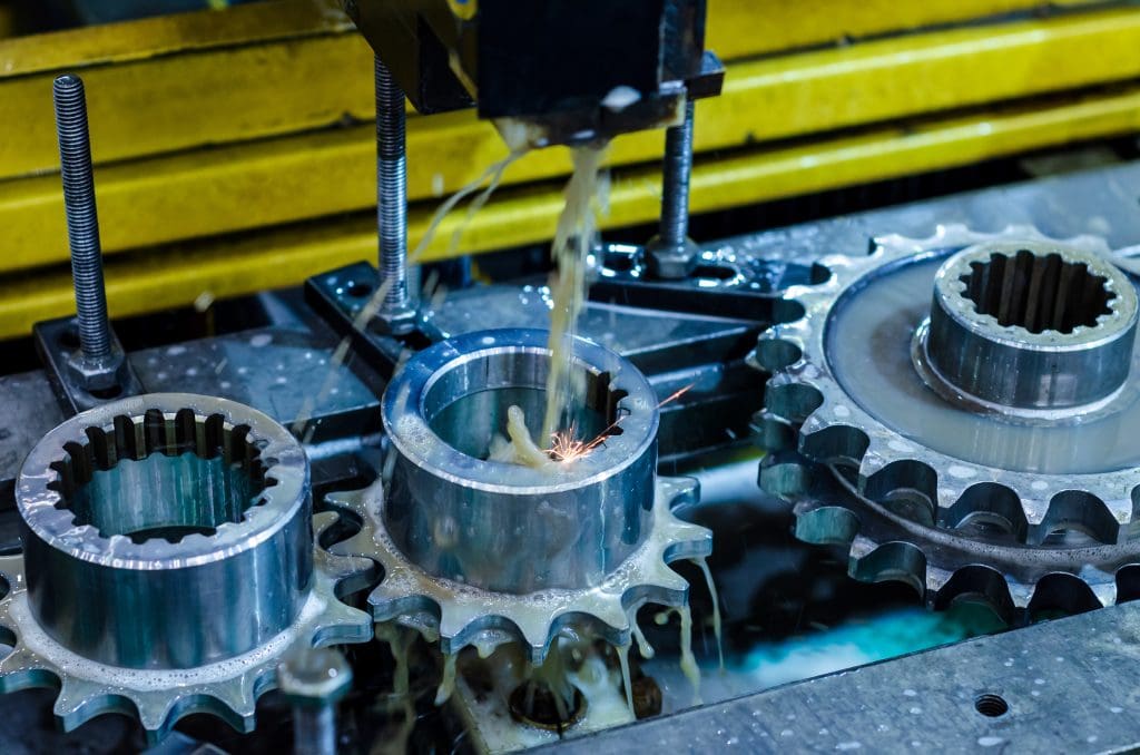 Close-up of three metal gears being machined, with coolant fluid and sparks visible as a tool works on the center gear; yellow industrial equipment is seen in the background.