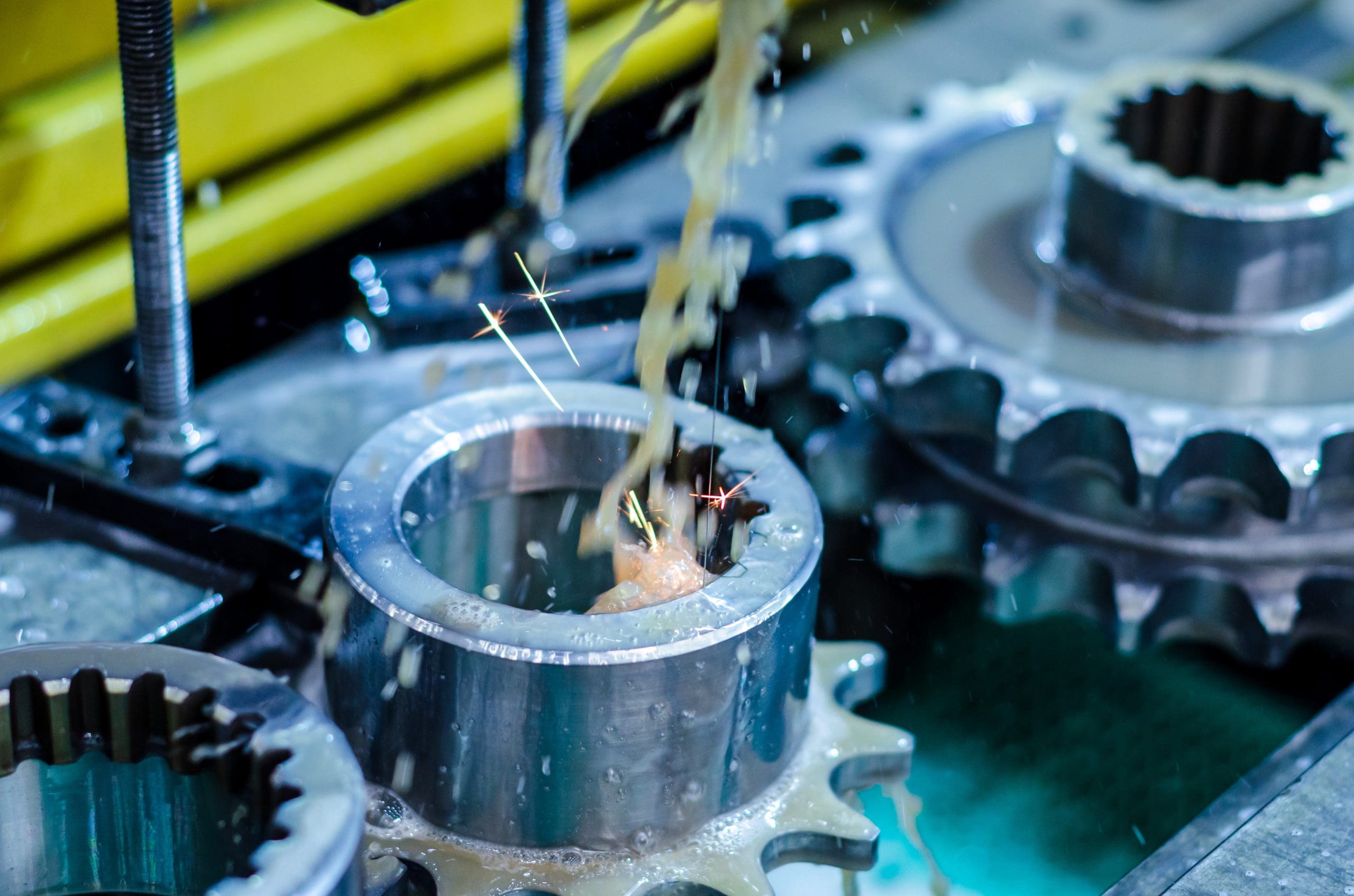 Close-up of a metal gear being machined with sparks and coolant fluid spraying, surrounded by other gears and industrial equipment in a factory setting.