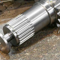 A large, shiny metal gear shaft with precision-cut teeth lies on a rough, speckled surface, likely in an industrial or workshop setting.