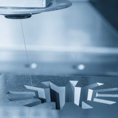 A wire electrical discharge machine cuts a precise, geometric pattern into a metal surface, with the thin wire visible above the workpiece. The scene has a cool, blue-toned lighting.