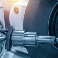 Close-up of a cylindrical metal workpiece being machined on an industrial lathe or grinding machine, with sharp, reflective surfaces and mechanical components visible.