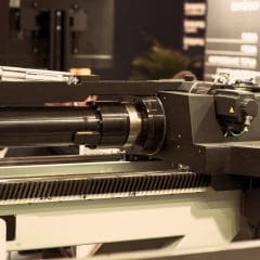 Close-up of a modern industrial CNC machine with metal components, gears, and an electric motor, used for precision manufacturing or machining in a factory setting. Background is blurred with some technical equipment visible.