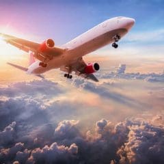 A commercial airplane flies above the clouds during sunset, with sunlight streaming across the sky and clouds below, creating a dramatic and colorful scene.