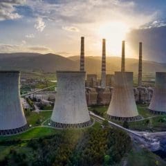 Aerial view of a power plant with four large cooling towers and three tall smokestacks at sunset, surrounded by greenery and mountains in the background.